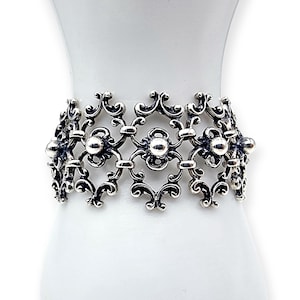 Könnte beinhalten: Verziertes Silberarmband mit filigranem Design. Das Armband weist ein sich wiederholendes Muster aus floralen und Rankenmotiven auf, die durch kleine, runde Perlen verbunden sind. Die aufwendigen Details und die polierte Oberfläche erzeugen eine Vintage- oder Antik-Ästhetik.