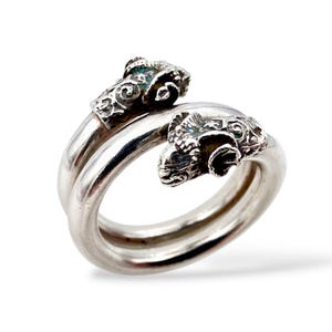 Könnte beinhalten: Ein silberner Ring mit zwei Widderkopfmotiven, die einander zugewandt sind. Der Ring hat ein Doppelband-Design. Die Widderköpfe sind aufwendig detailliert. Der Ring wird vor einem weißen Hintergrund präsentiert, der seinen metallischen Glanz hervorhebt.
