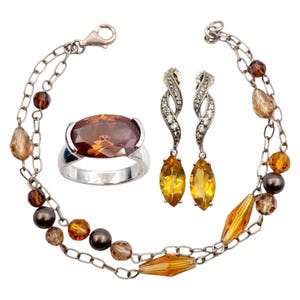 Könnte beinhalten: Ein Schmuckset mit einem silbernen Ring mit einem großen, ovalen, bernsteinfarbenen Stein, einem silbernen Armband mit Bernstein- und braunen Perlen und einem Paar silberner Ohrringe mit gelben tropfenförmigen Steinen und klaren Akzenten.