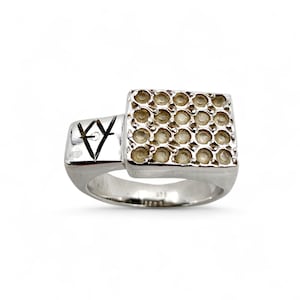 Könnte beinhalten: Ein silberner Ring mit quadratischer Oberseite, die mit einem Gitter aus kleinen, runden, hellfarbigen Steinen besetzt ist. Der Ring hat ein einzigartiges Design mit einem geometrischen Symbol an der Seite.