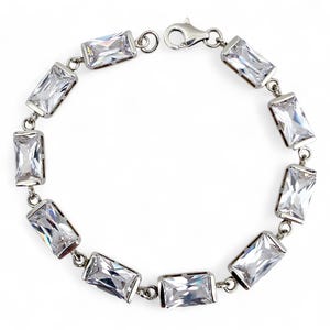 Könnte beinhalten: Ein silbernes Armband mit rechteckigen, klaren Edelsteinen. Die Edelsteine sind in einem silbernen Metallrahmen gefasst und miteinander verbunden. Das Armband hat einen Karabinerverschluss. Der Schmuck wird auf weißem Hintergrund präsentiert.