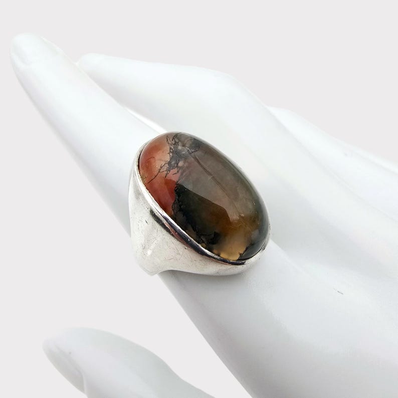 Puede incluir: Un anillo de plata con una gran piedra preciosa de forma ovalada. La piedra preciosa es una mezcla de colores marr&oacute;n, naranja y verde con vetas negras.