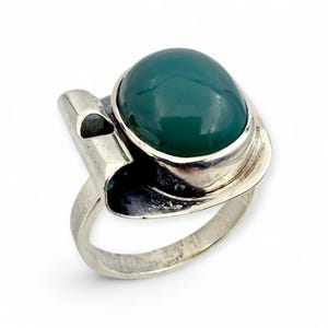 Könnte beinhalten: Silberring mit einem großen, ovalen, dunkelgrünen Edelstein. Der Ring hat ein ungewöhnliches, asymmetrisches Design mit einem gebogenen Band und einem dekorativen Element. Der Edelstein ist in einer polierten Silberfassung gefasst.