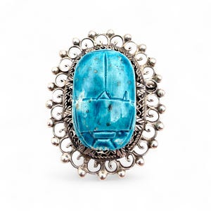 Könnte beinhalten: Ovaler silberfarbener Ring mit einem türkisblauen Skarabäus-Design. Der Käfer ist leuchtend türkisblau mit geschnitzten Details. Der Ring hat einen verzierten, durchbrochenen Rand mit einem dekorativen Muster.