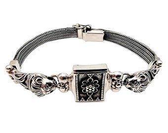 May include: A silver bracelet with a woven chain design. The bracelet features lion head accents on each end and a central square element with a floral pattern. The clasp is rectangular.