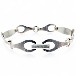 Könnte beinhalten: Ein silberfarbenes Armband mit einer Reihe von länglichen, oval geformten Gliedern, die durch kleinere, kreisförmige Glieder verbunden sind. Das Armband hat einen rechteckigen Verschluss. Der Schmuck ist vor weißem Hintergrund dargestellt.