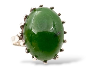 May include: A silver ring featuring a large, oval-shaped, glossy green stone. The stone is surrounded by small, silver, rounded prongs. The ring band is a simple, polished silver.