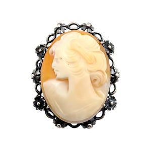 Könnte beinhalten: Ovaler Cameo-Anhänger mit dem Profil einer Frau mit fließendem Haar, geschnitzt in Creme- und Orangetönen. Der Cameo ist in einem silberfarbenen Rahmen mit floralen Akzenten gefasst und erzeugt eine Vintage-Ästhetik.