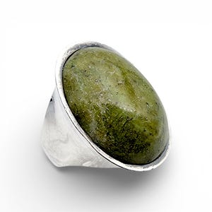 May include: A silver ring with a large, oval green stone. The stone has a mottled appearance with different shades of green. The ring's band is wide and made of polished silver, creating a bold, modern look. The ring is set against a white background.