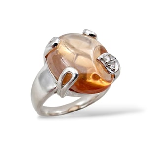 May include: A silver ring with a large, oval amber gemstone. The gemstone is set with small diamonds and a silver band. The ring has a modern, elegant design. The ring is suitable for everyday wear or special occasions.
