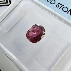 Può includere: Una pietra preziosa ovale rosa in un contenitore di plastica trasparente.