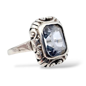 Könnte beinhalten: Ein silberner Ring mit einem rechteckigen, hellblauen Edelstein. Der Ring hat ein verziertes, gerolltes Design um den Edelstein und am Band. Der Ring ist vor einem weißen Hintergrund platziert.