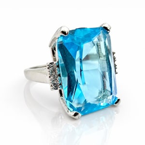 Könnte beinhalten: Ein silberfarbener Ring mit einem großen, rechteckigen, facettierten leuchtend blauen Edelstein als Mittelstück. Die Ringschiene ist auf beiden Seiten der zentralen Fassung mit drei kleinen, klaren, runden Steinen besetzt und vier Krappen.