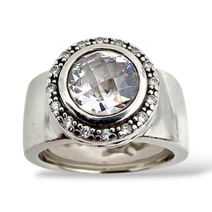 Puede incluir: Un anillo de plata con una gran piedra preciosa clara y facetada en el centro, rodeada por un halo de piedras claras más pequeñas. El anillo tiene una banda ancha y un acabado pulido, creando un efecto brillante.