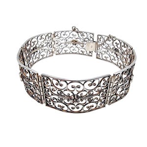 Könnte beinhalten: Ein silbernes Filigranarmband mit einem kunstvollen, durchbrochenen Design. Das Armband besteht aus rechteckigen Elementen, die durch Scharniere verbunden sind, mit einem floralen Motiv in der Mitte. Der Verschluss ist sichtbar. Auf weißem Hintergrund.