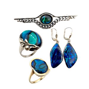 Könnte beinhalten: Eine Schmuckkollektion mit schillernden blau-grünen Abalone-Muscheln. Das Set umfasst eine silberne Brosche, zwei Ringe und ein Paar Ohrringe. Die Ohrringe und Ringe haben silberne und goldene Fassungen.