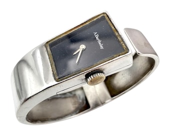 May include: A silver wristwatch with a rectangular black face. The watch has a silver band and a gold-coloured crown. The watch face displays the text "A. Barthelay".