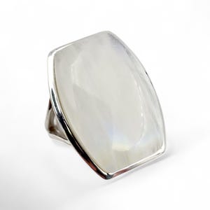 Könnte beinhalten: Ein auffälliger Ring mit einem großen, rechteckigen, irisierenden weißen Edelstein, der in einem silbernen Band gefasst ist. Der Edelstein hat einen subtilen, perlmuttartigen Glanz. Das Design des Rings ist modern und kühn.