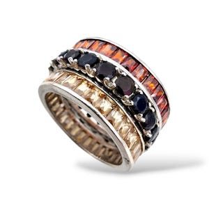 Könnte beinhalten: Ein Stapel von drei silbernen Ringen mit farbigen Edelsteinen. Der oberste Ring hat rechteckige orangefarbene Steine, der mittlere Ring runde dunkelblaue Steine und der untere Ring rechteckige hellbraune Steine. Vor weißem Hintergrund.