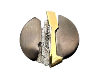 Könnte beinhalten: Silber- und goldfarbene Brosche mit abstraktem Design. Die Brosche hat eine runde Form mit zwei gebürsteten Silberabschnitten und einem zentralen goldfarbenen Element. Ein Abschnitt ist mit kleinen, funkelnden Steinen besetzt.