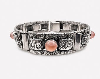 Puede incluir: Una pulsera de metal plateado con una superficie rugosa y muy texturizada con eslabones rectangulares. La pulsera está acentuada con piedras cabujón lisas, redondas y de color rosa pálido colocadas a intervalos a lo largo del centro.