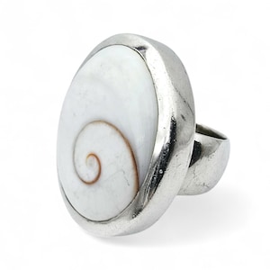 Pode incluir: Anel de prata com uma grande concha branca ovalada com um padrão em espiral. A concha é fixada em uma grossa faixa de prata polida. O anel tem um design arrojado.