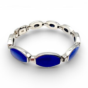 Könnte beinhalten: Ein silbernes Armband mit ovalen, tiefblauen Einsätzen. Das Armband besteht aus verbundenen Segmenten, jedes mit einem polierten Silberrahmen und einem leuchtend blauen Zentrum. Der Verschluss ist sichtbar, und das Armband ist vor einem weißen Hintergrund platziert.