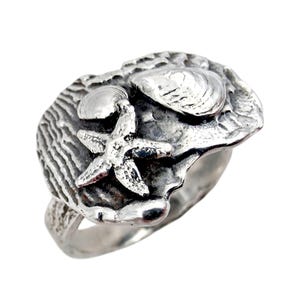 Könnte beinhalten: Silberner Ring mit Muscheldesign, einschließlich eines Seesterns und zweier Muscheln. Der Ring hat eine strukturierte Oberfläche und ein poliertes Finish, ideal für Strandliebhaber.