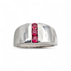Könnte beinhalten: Ein silberner Ring mit drei leuchtend roten Edelsteinen. Der Ring hat ein breites, gebogenes Band mit strukturierter Oberfläche. Die Edelsteine sind rund und gleichmäßig angeordnet und verleihen dem Design einen Farbtupfer. Der Ring ist etwa 2 cm breit.