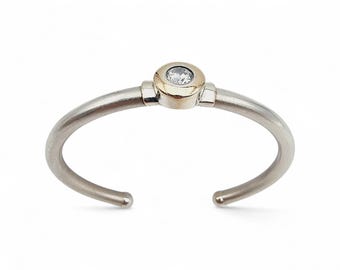 Op de afbeelding: Een zilverkleurige manchetarmband met een goudkleurig middenstuk. Het middenstuk heeft een kleine, ronde, heldere edelsteen. De armband heeft een eenvoudig, elegant ontwerp, ideaal voor dagelijks gebruik.
