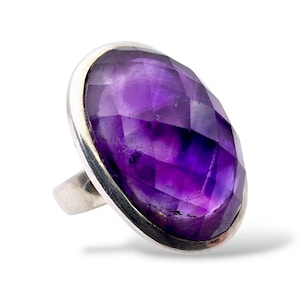 May include: A silver ring with a large, oval-cut amethyst gemstone. The vibrant purple stone is set in a silver band. The ring's design is simple, highlighting the gemstone's colour and facets. The ring is a statement piece.