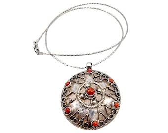 Puede incluir: Un collar con colgante de plata con un diseño circular. El colgante presenta un diseño central con detalles de coral rojo y un borde decorativo. El collar tiene una delicada cadena de plata.