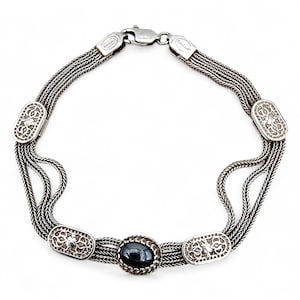 Könnte beinhalten: Silberarmband mit geflochtenem Kettendesign und dekorativen ovalen Gliedern. Das Armband hat einen zentralen dunklen Stein und einen Verschluss. Ein Schmuckstück für den täglichen Gebrauch.
