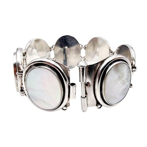 Könnte beinhalten: Ein silberfarbenes Armband mit ovalen Perlmutt-Inlays. Das Armband besteht aus mehreren verbundenen Segmenten, jedes mit einem ovalen Inlay. Der Verschluss ist geöffnet und zeigt den Scharniermechanismus.
