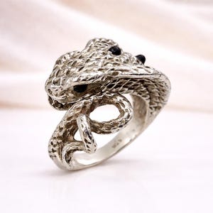 Könnte beinhalten: Ein silberner Ring in Form einer Schlange. Der Schlangenkopf ist mit strukturierten Schuppen und zwei schwarzen Edelsteinaugen versehen. Der Körper der Schlange windet sich um den Finger und erzeugt ein einzigartiges Design.