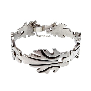 Könnte beinhalten: Ein silberfarbenes Armband mit Blattmuster. Das Armband besteht aus miteinander verbundenen, blattförmigen Gliedern, mit einem sicheren Verschluss und einer Sicherheitskette. Die Blätter haben detaillierte Adern. Dieses Schmuckstück ist für den Alltag geeignet, ca. 18 cm lang.