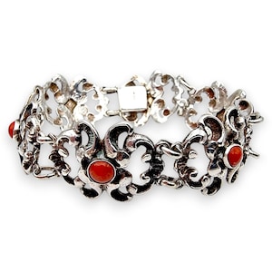 Könnte beinhalten: Silberfarbenes Armband mit einem verzierten, durchbrochenen Design. Das Armband hat eine Reihe von stilisierten Blumenformen, die jeweils mit einem kleinen, runden, roten Stein akzentuiert sind. Der Verschluss ist ein einfaches, rechteckiges Design. Schmuck.