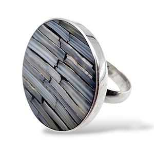 Könnte beinhalten: Silberring mit einem großen, ovalen Ringkopf. Der Ringkopf ist mit einem Mosaik aus grauen und weißen rechteckigen Teilen verziert, das ein strukturiertes Muster bildet. Der Ring hat einen schlichten, polierten Ring.