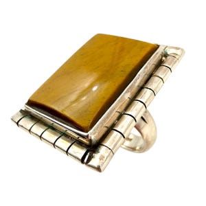 Op de afbeelding: Een zilveren ring met een grote, rechthoekige tijgeroogsteen. De steen heeft een warme, goudbruine kleur. De ring heeft een uniek ontwerp met een reeks kleine, rechthoekige zilveren staven aan de zijkanten van de steen.
