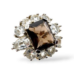 May include: A silver ring with a central rectangular, smoky brown gemstone, encircled by smaller, clear, round and pear-shaped stones. The ring's design is set against a white backdrop, accentuating the sparkle of the gemstones.