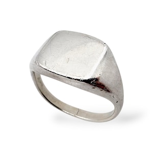 May include: A silver signet ring with a square face. The ring has a polished finish and a simple, classic design. The ring is likely made of sterling silver or a similar metal.