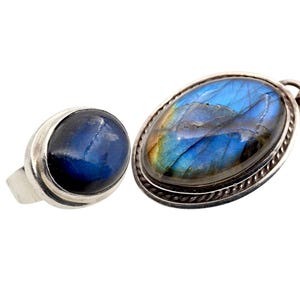 Könnte beinhalten: Ein silbernes Ring- und Anhänger-Halsketten-Set. Der Ring hat einen runden, dunkelblauen Edelstein. Der Anhänger hat einen ovalen, irisierenden blauen Edelstein mit silbernem Seilrand. Die Halskette hat eine Silberkette.