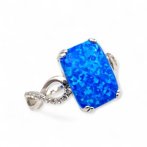 Könnte beinhalten: Ein silberner Ring mit einem großen, rechteckigen, leuchtend blauen Opal-Edelstein. Der Opal hat ein gesprenkeltes Aussehen. Das Ringband hat ein Unendlichkeitszeichen-Design, verziert mit kleinen, klaren Edelsteinen. Die Fassung um den Opal ist ebenfalls silberfarben.