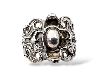Puede incluir: Anillo de plata ornamentado con un diseño ovalado central y elaborados adornos. El anillo tiene un aspecto vintage y antiguo, con un acabado deslustrado. El anillo es una pieza llamativa, adecuada para añadir un toque de elegancia.