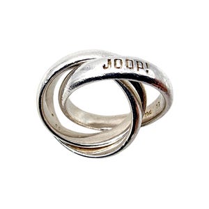 Könnte beinhalten: Ein silberfarbener Metallring bestehend aus drei ineinander verschlungenen Bändern. Das obere Band zeigt den geprägten Schriftzug JOOP! in Großbuchstaben. Die Metalloberfläche wirkt leicht texturiert und abgenutzt mit sichtbaren Markierungen an den inneren Bändern.