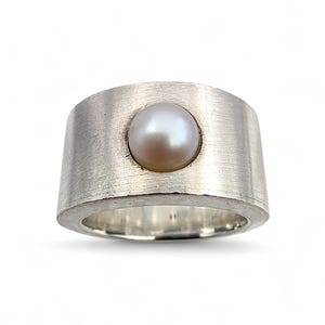 Könnte beinhalten: Ein breiter, silberfarbener Ring mit gebürsteter Metalloberfläche. Der Ring hat eine große, runde, cremefarbene Perle in der Mitte. Das Band ist dick und leicht ausgestellt, was einen kühnen und modernen Look erzeugt. Es ist ein Schmuckstück.