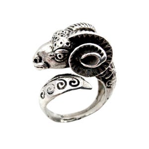 Könnte beinhalten: Ein silberner Ring mit einem Widderkopf-Design. Der Widderkopf hat detaillierte Merkmale, einschließlich Hörner und Gesichtszüge. Das Ringband hat ein dekoratives Wirbelmuster. Der Ring ist vor einem weißen Hintergrund.