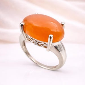 Könnte beinhalten: Ein silberner Ring mit einem großen, ovalen, orangefarbenen Edelstein. Der Ring hat ein detailliertes filigranes Design am Band und einen kleinen Akzentstein. Der Edelstein ist hoch gesetzt und zeigt seine leuchtende Farbe und glatte Oberfläche.