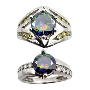 Könnte beinhalten: Zwei silberne Ringe mit großen, schillernden, runden Edelsteinen. Ein Ring hat ein geometrisches Design mit kleineren grünen Steinen. Der andere Ring hat ein Band mit kleinen, klaren Steinen. Beide Ringe sind vor weißem Hintergrund.