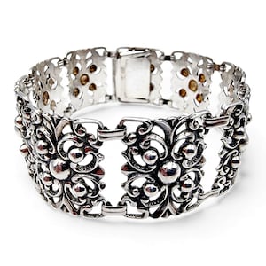 Könnte beinhalten: Ein silbernes Armband mit einem verzierten floralen Design. Das Armband weist detaillierte Schnörkel und kleine, kugelförmige Akzente auf. Der Verschluss ist rechteckig und das Armband ist vor einem weißen Hintergrund platziert.